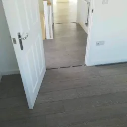 Świeżo ułożone panele podłogowe w odcieniach szarości, widoczne otwarte białe drzwi z chromowaną klamką, przejście do dalszej części domu z grzejnikiem na ścianie.