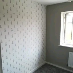 Pokój po remoncie: ściana pokryta białą tapetą z szarym wzorem królików, sąsiadująca z szarą ścianą pomalowaną farbą; szary dywan na podłodze, biała listwa przypodłogowa, okno z białymi ramami...