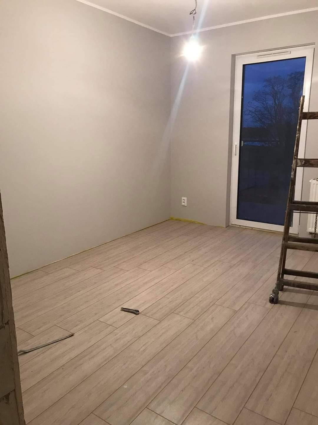 Pokój w trakcie remontu z ułożonymi panelami podłogowymi, drabina oparta o ścianę przy oknie, widoczne narzędzia na podłodze, ściany pomalowane na szaro.