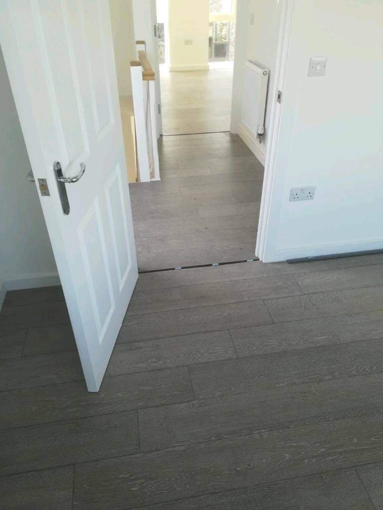 Świeżo ułożone panele podłogowe w odcieniach szarości, widoczne otwarte białe drzwi z chromowaną klamką, przejście do dalszej części domu z grzejnikiem na ścianie.