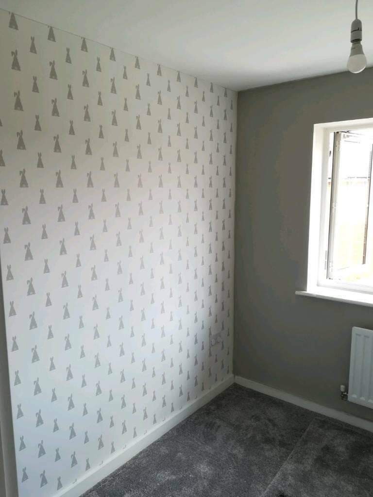 Pokój po remoncie: ściana pokryta białą tapetą z szarym wzorem królików, sąsiadująca z szarą ścianą pomalowaną farbą; szary dywan na podłodze, biała listwa przypodłogowa, okno z białymi ramami...