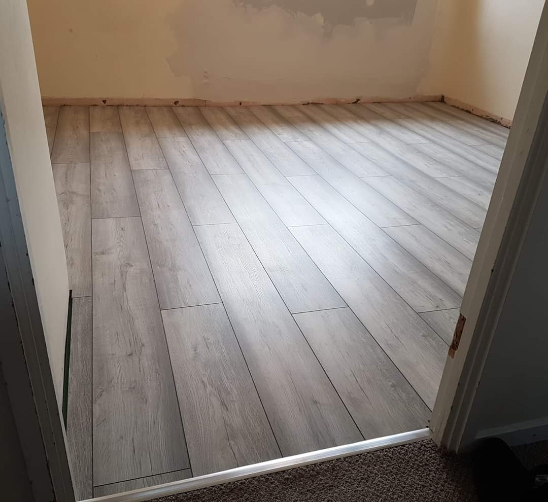 Świeżo ułożone panele podłogowe w odcieniach szarości z widoczną listwą przypodłogową i surowymi ścianami przed malowaniem, widok z progu drzwi.