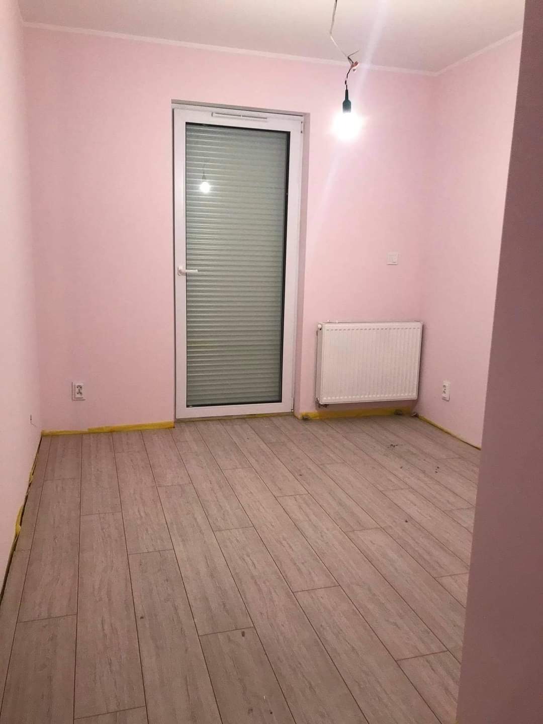 Pokój po remoncie: świeżo ułożone panele podłogowe w odcieniach beżu, różowe ściany zabezpieczone taśmą malarską przy podłodze, białe drzwi balkonowe z żaluzjami, grzejnik i wisząca żarówka.