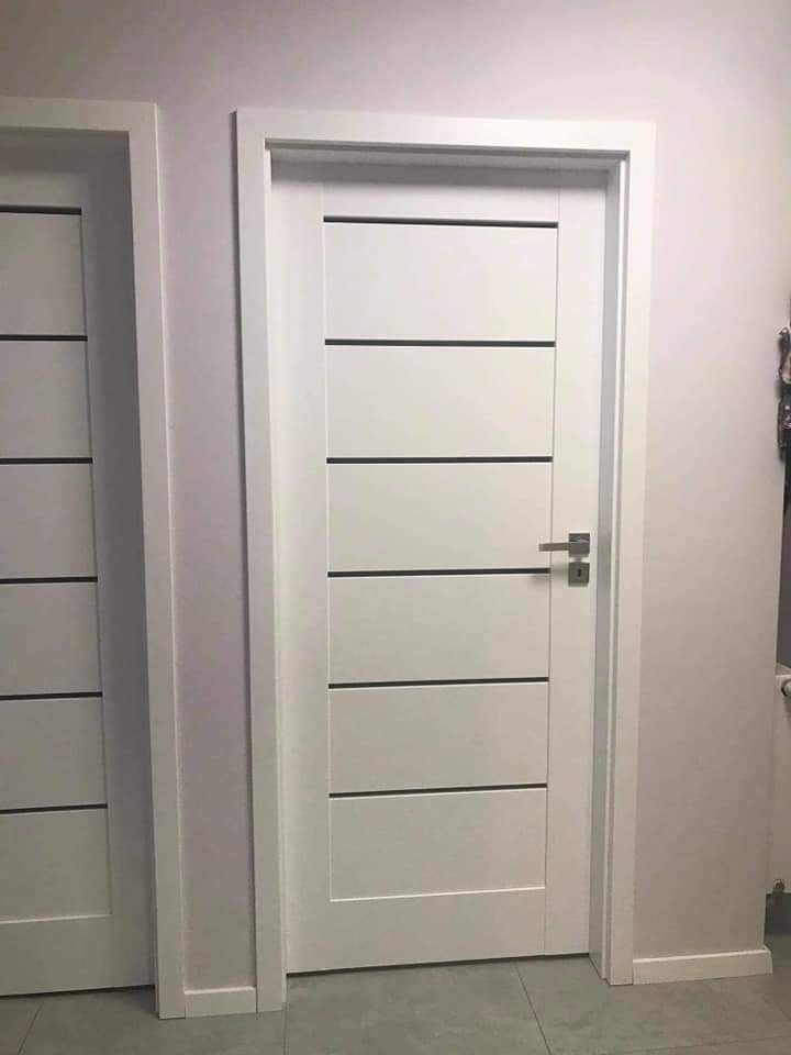 Białe drzwi wewnętrzne z poziomymi, czarnymi wstawkami, zamontowane w ościeżnicy, widoczne obok drugie, podobne drzwi.