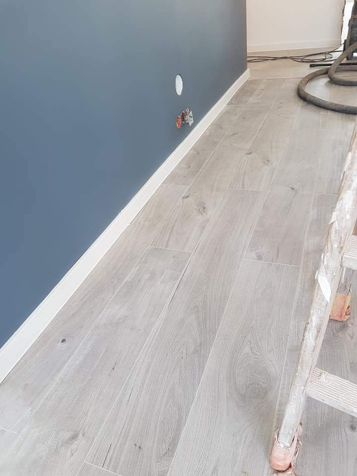 Jasnoszare panele podłogowe ułożone wzdłuż ciemnoniebieskiej ściany z białym cokołem, widoczna drabina malarska i rury od odkurzacza przemysłowego.