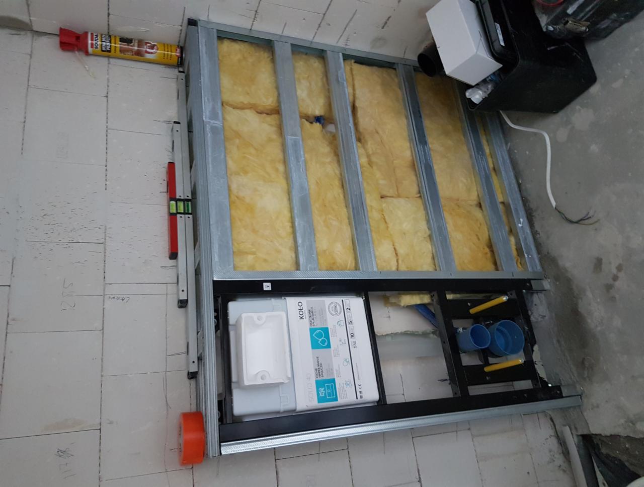 Metalowa konstrukcja zabudowy z izolacją z wełny mineralnej, widoczny stelaż podtynkowy WC marki Koło, poziomica oraz ślady ołówka na podłodze.