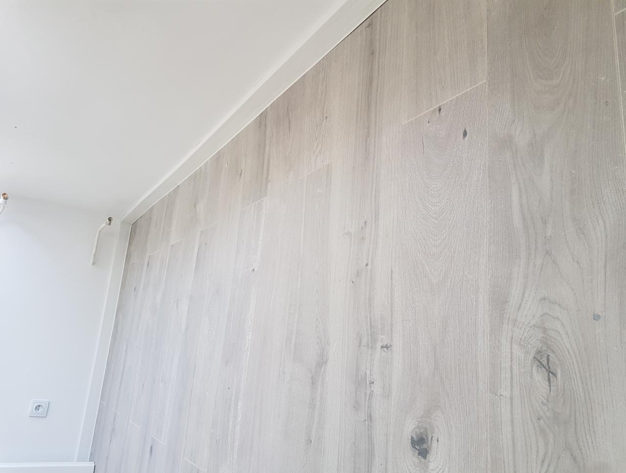 Ściana wykończona panelami podłogowymi w odcieniach szarości, widoczna listwa przypodłogowa i gniazdko elektryczne na białej ścianie obok.