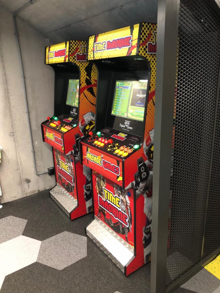 Dwa automaty do gier arcade 'Time Machine' stojące obok siebie, obudowy w kolorze czerwonym i żółtym, widoczne ekrany z grafiką gry, umieszczone w pomieszczeniu z betonową ścianą i wykładziną...