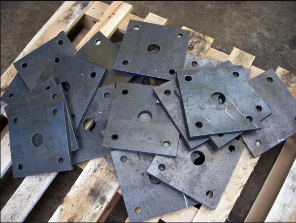 Stos szarych, kwadratowych, metalowych płyt z centralnym otworem i czterema mniejszymi otworami na rogach, ułożonych na drewnianej palecie.