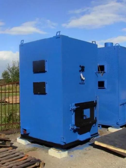 Dwa niebieskie, metalowe kotły przemysłowe umieszczone na zewnątrz, z widocznymi czarnymi drzwiczkami i elementami mocującymi, ustawione na betonowych podstawach, na tle pochmurnego nieba.