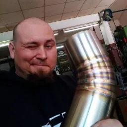 Uśmiechnięty mężczyzna z brodą mruga okiem, prezentując spawany element rury ze stali nierdzewnej z widocznymi, kolorowymi przebarwieniami po spawaniu TIG, na tle warsztatu.