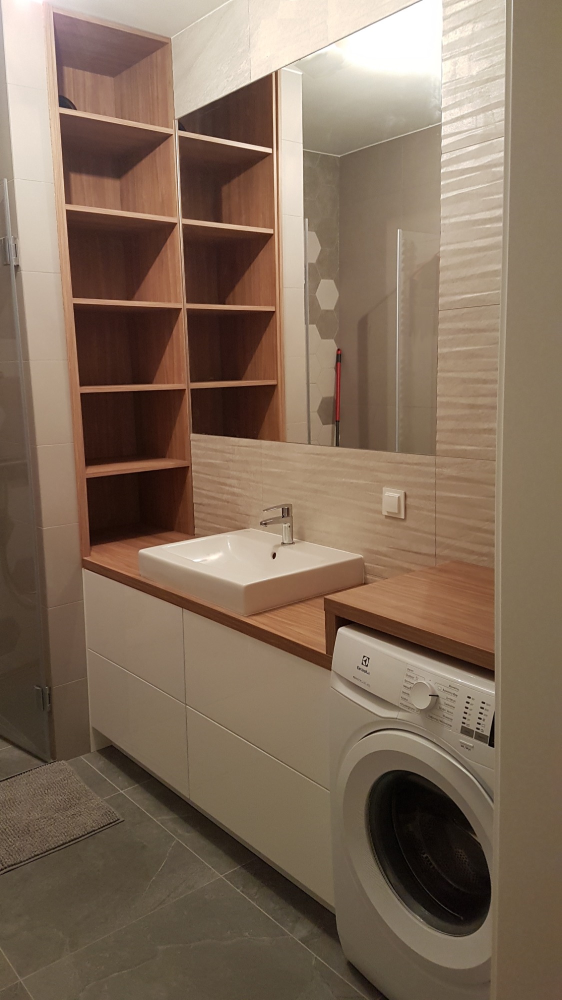 Nowoczesna łazienka z drewnianym blatem, białą umywalką nablatową, szafkami pod umywalką, regałem z drewnianymi półkami i pralką Electrolux.