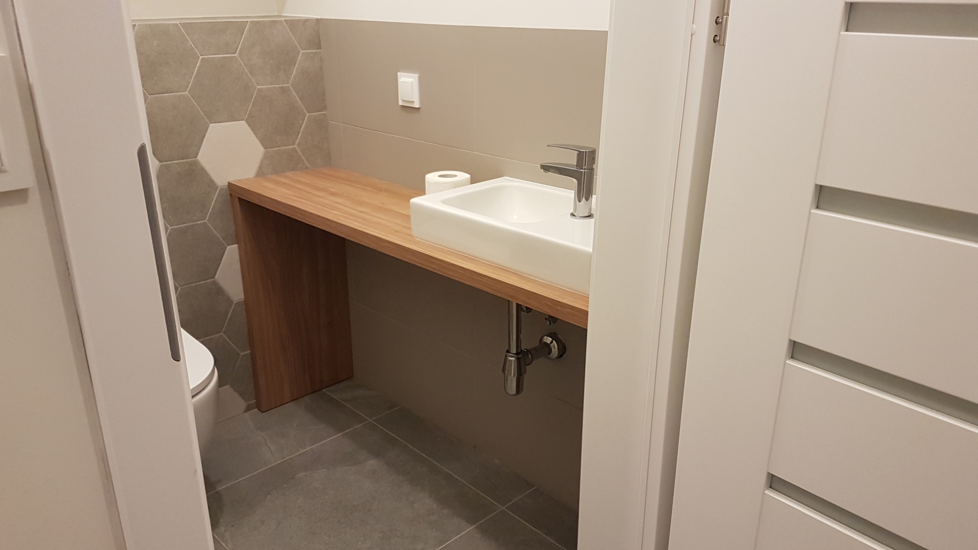 Nowoczesna łazienka z umywalką nablatową na drewnianym blacie, heksagonalne płytki na ścianie i białe drzwi z poziomymi przeszkleniami.