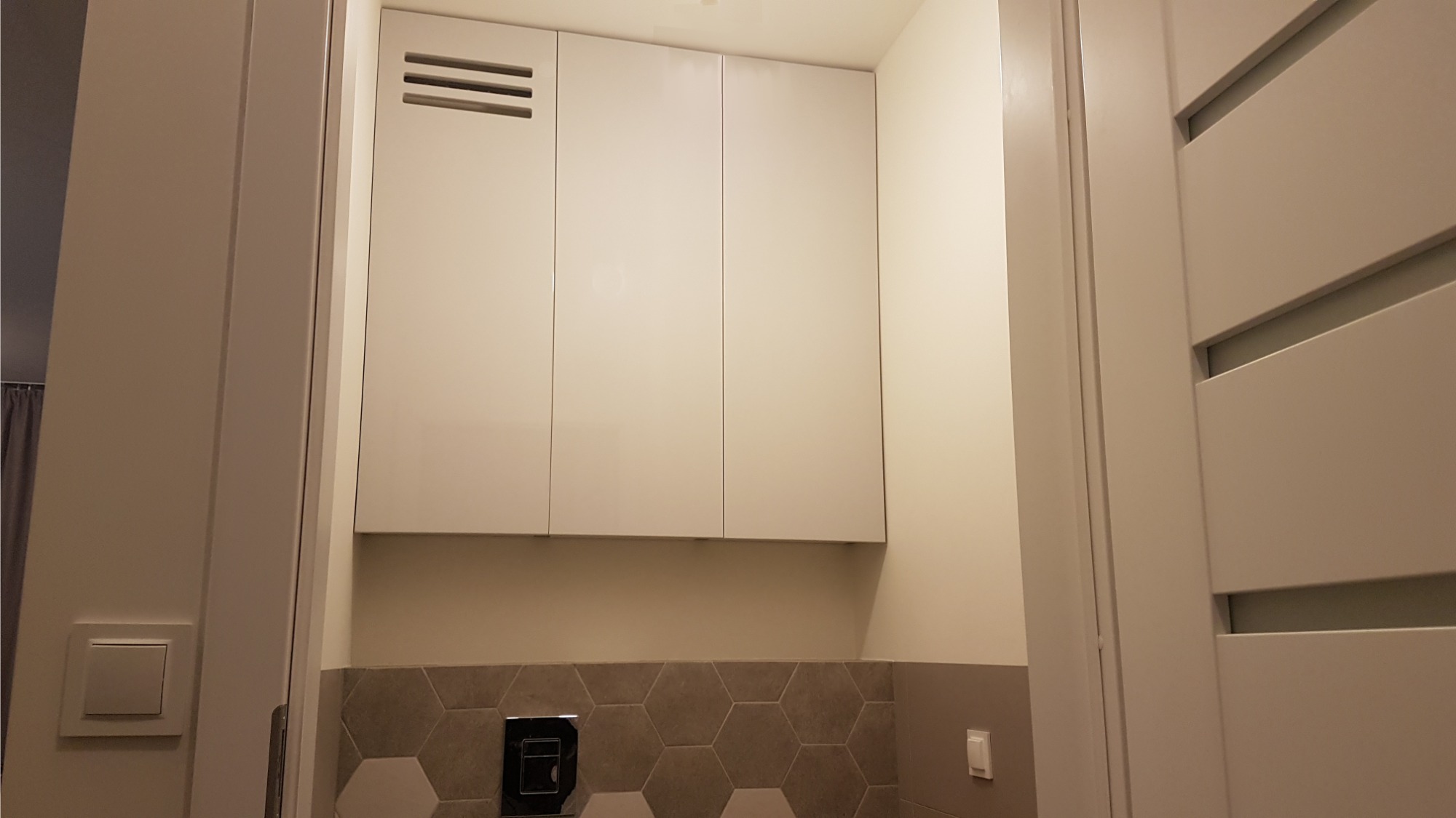 Białe szafki wiszące w toalecie nad zabudowaną spłuczką, heksagonalne płytki na ścianie, widok zza uchylonych drzwi.