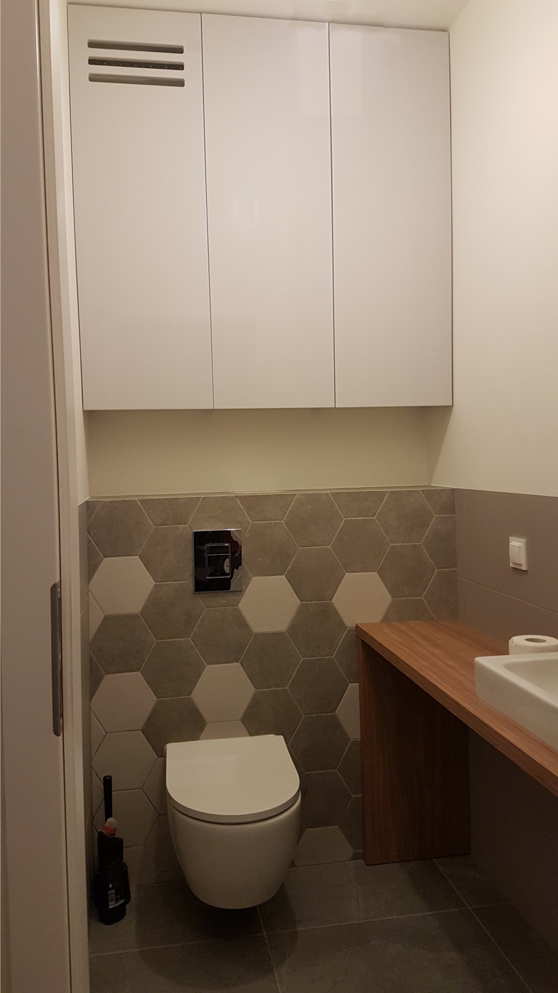 Nowoczesna łazienka z wiszącą toaletą, heksagonalnymi płytkami w odcieniach szarości i białymi szafkami wiszącymi z poziomymi szczelinami wentylacyjnymi, drewniany blat z umywalką.