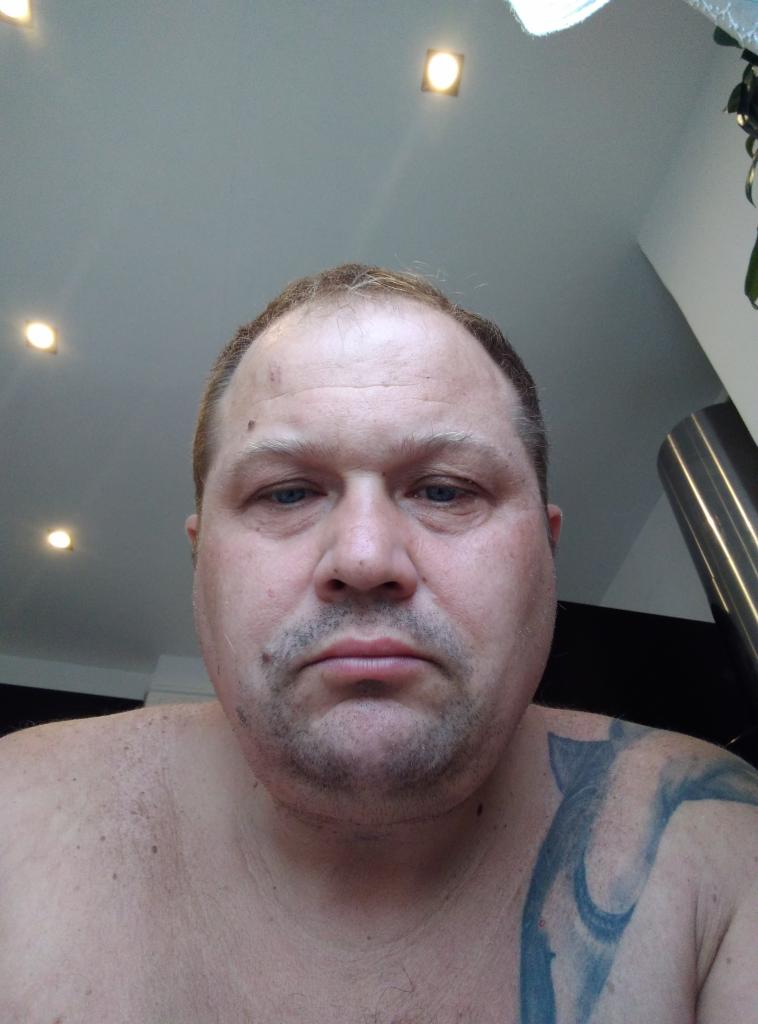 Autoportret mężczyzny z tatuażem na ramieniu, patrzącego w dół na aparat, pod sufitem z wbudowanymi lampami.