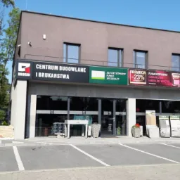 Fasada budynku Centrum Budowlanego i Brukarstwa z szyldami reklamowymi, ekspozycją płyt brukowych Libet i parkingiem.