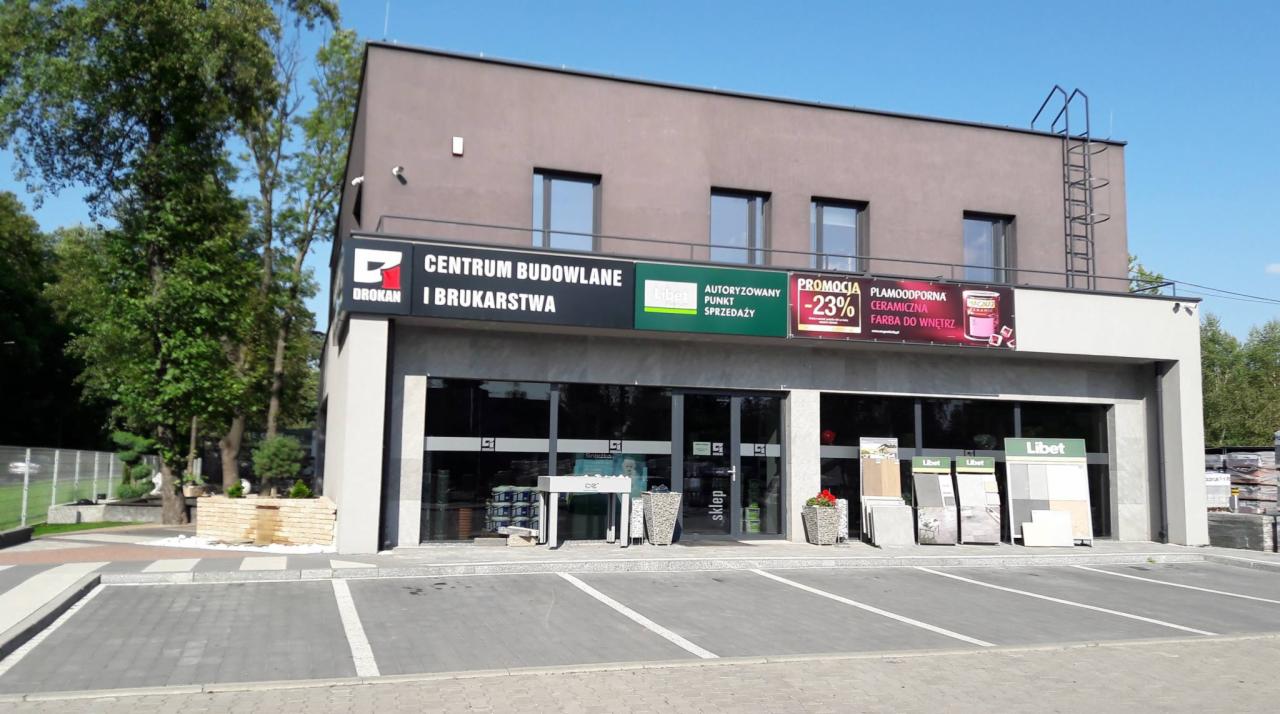 Fasada budynku Centrum Budowlanego i Brukarstwa z szyldami reklamowymi, ekspozycją płyt brukowych Libet i parkingiem.