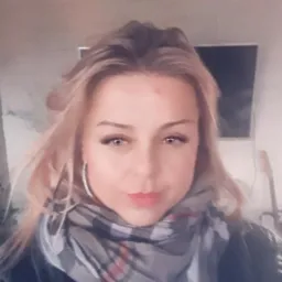 Portret kobiety z blond włosami, szarym szalem i czarnej kurtce na tle jasnej ściany i telewizora, ujęcie z bliska, lekko od dołu.