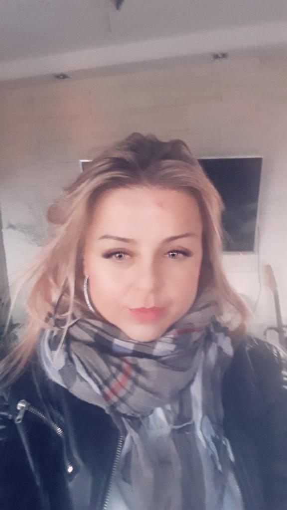 Portret kobiety z blond włosami, szarym szalem i czarnej kurtce na tle jasnej ściany i telewizora, ujęcie z bliska, lekko od dołu.