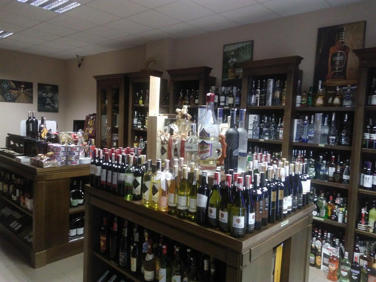 Wnętrze sklepu z alkoholem, półki i stoły ekspozycyjne wypełnione butelkami wina, wódki i innych trunków, dekoracyjne opakowania prezentowe.