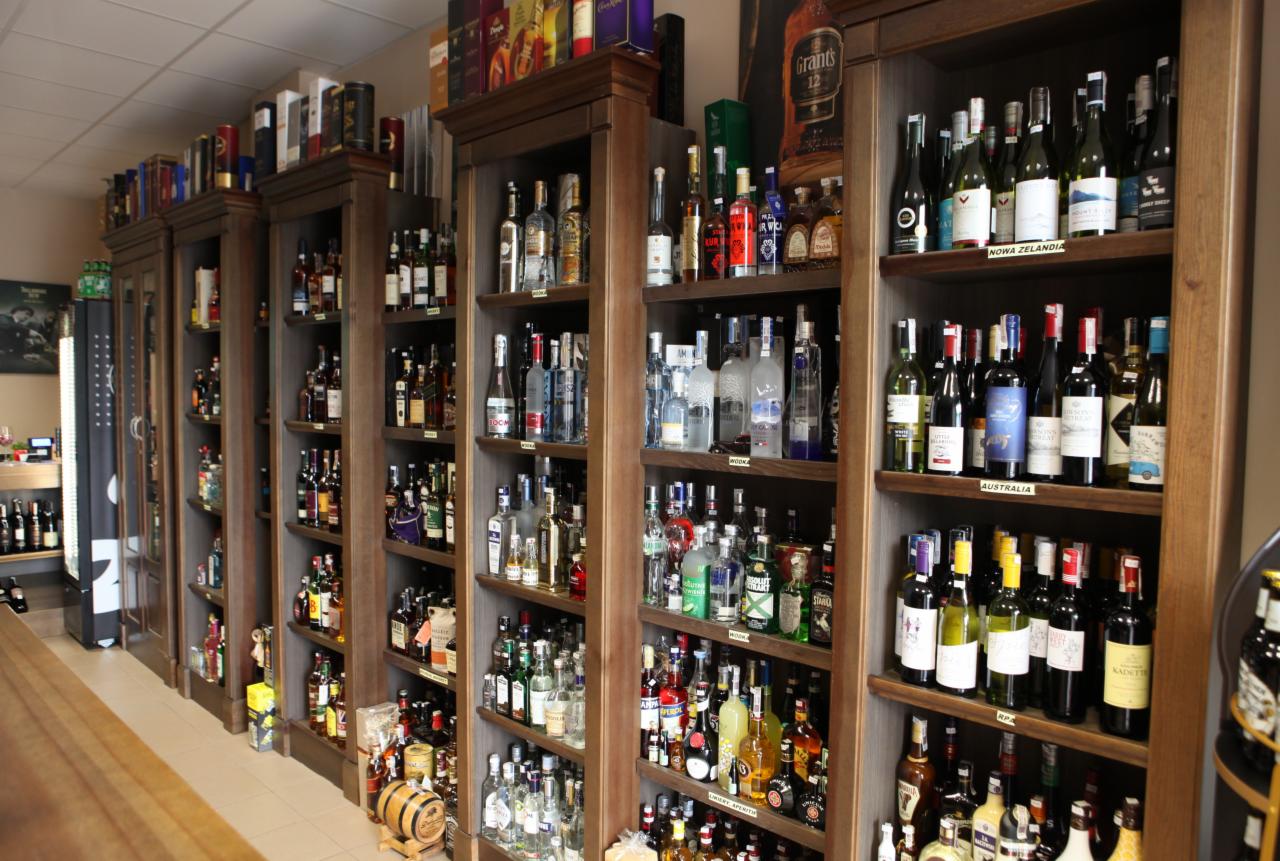 Pełne drewniane regały sklepowe z różnorodnymi butelkami alkoholi, w tym wina z Nowej Zelandii i Australii, oraz whisky Grant's 12, stojące w rzędzie w jasnym wnętrzu.