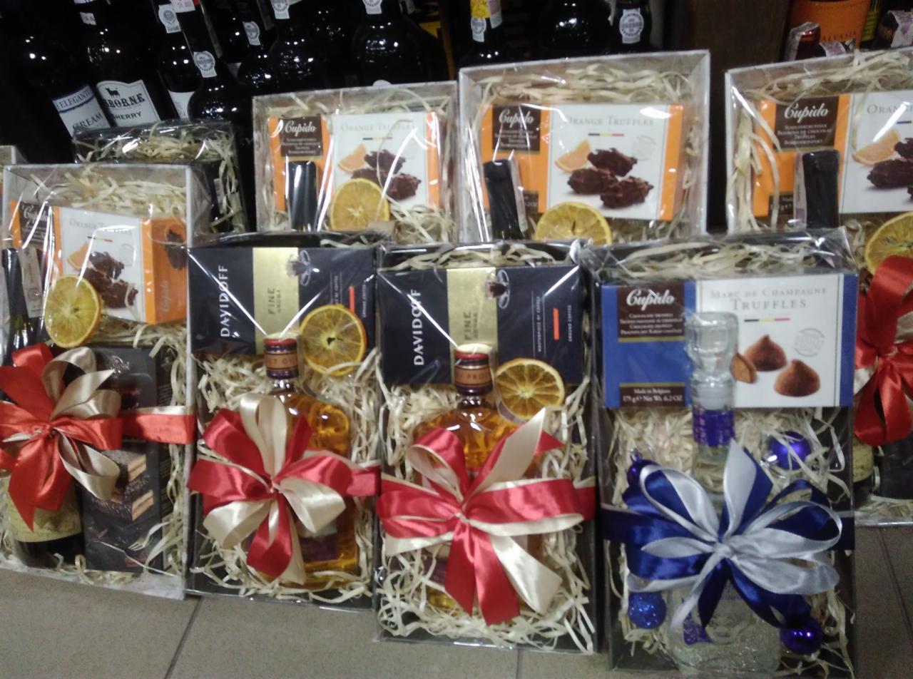 Ustawione w rzędzie zapakowane zestawy upominkowe zawierające butelki alkoholu, czekolady i suszone pomarańcze, owinięte w celofan i ozdobione wstążkami w kolorach czerwieni, bieli i błękitu.