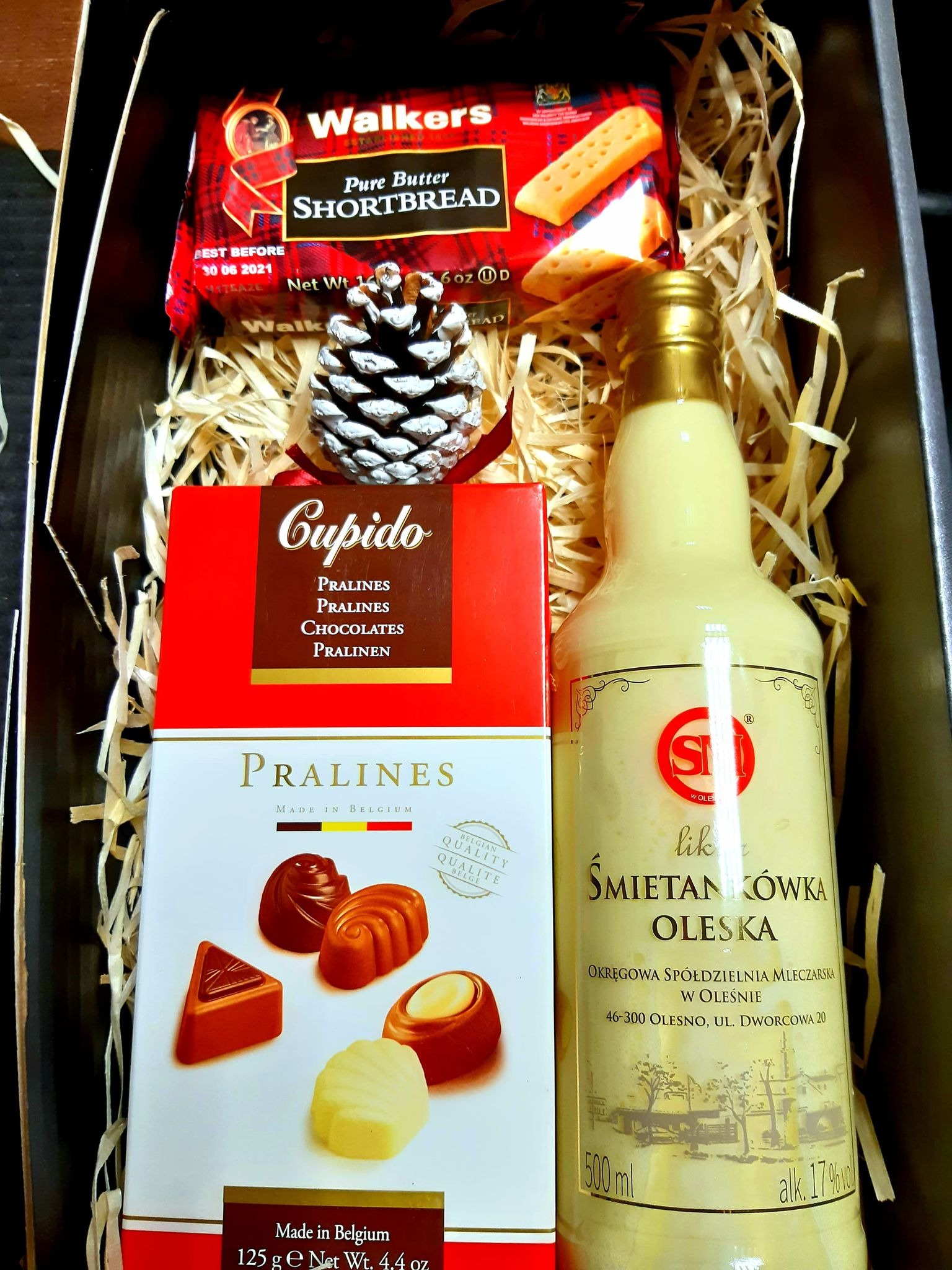 Zestaw upominkowy zawierający butelkę likieru Śmietankówka Oleska, pudełka pralin Cupido i Belgian Pralines oraz szkockie ciasteczka Walkers Shortbread, udekorowany szyszką i siankiem w ciemnym...