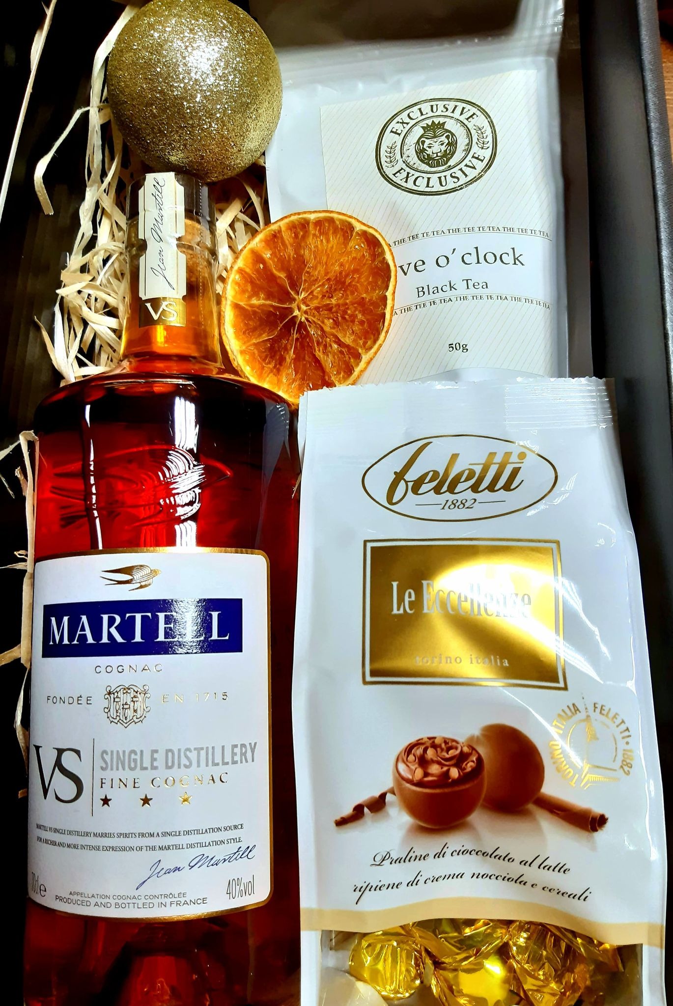 Zestaw upominkowy zawierający butelkę koniaku Martell VS Single Distillery, herbatę Exclusive i praliny Feletti, uzupełniony suszoną pomarańczą i złotą bombką, ułożony w pudełku wypełnionym...