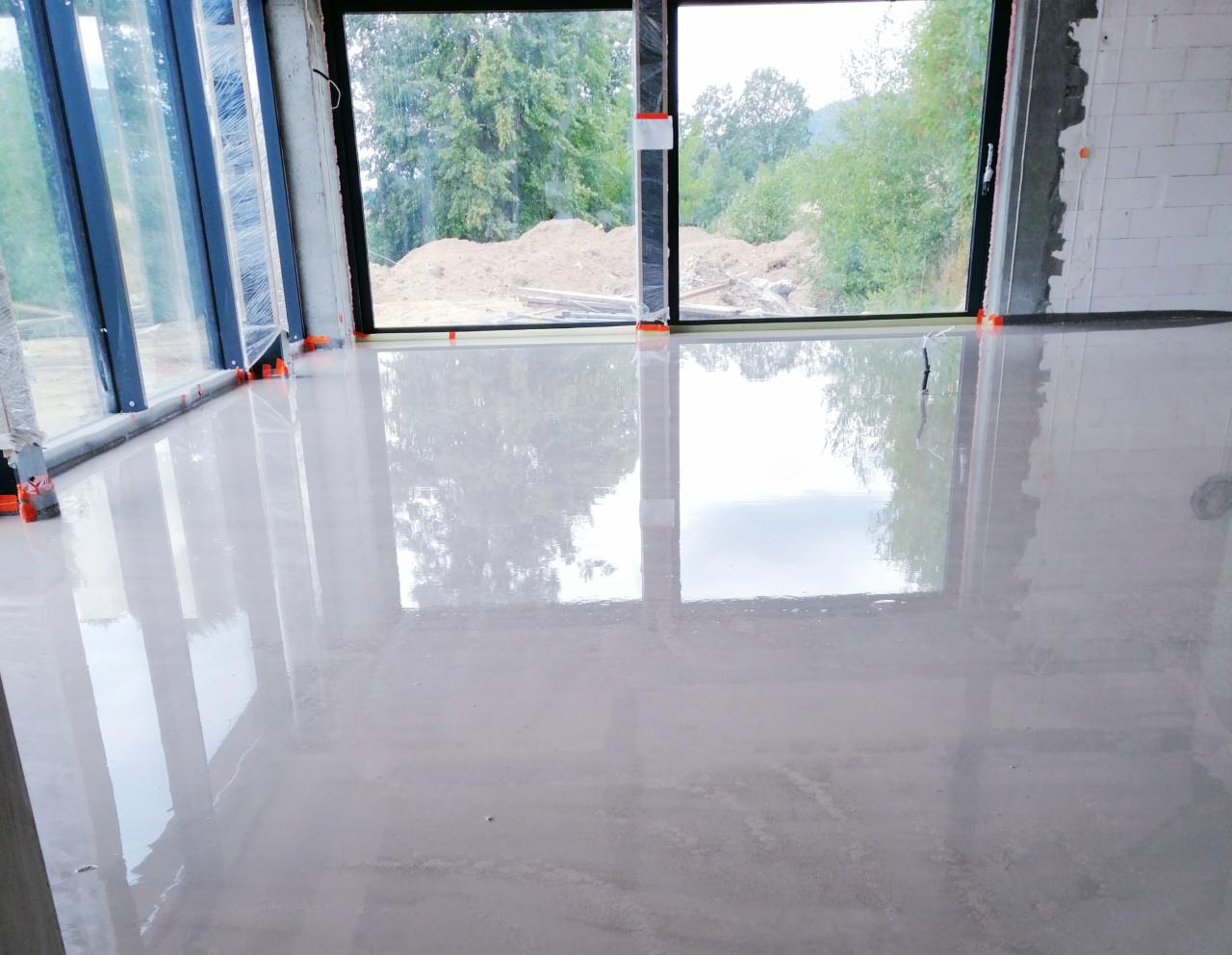 Świeżo wylana, gładka wylewka anhydrytowa z widocznymi refleksami światła, pomarańczowe znaczniki poziomowania przy oknach z widokiem na zewnątrz, surowe ściany z betonu komórkowego.