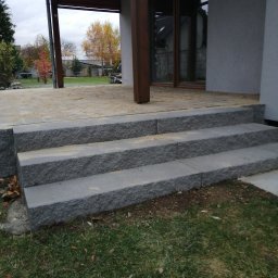 adamex-bruk - Granitowe schody wejściowe do domu z tarasem, widok z dołu. Szara kostka granitowa, jesienne drzewa w tle. Detal wykończenia posesji.