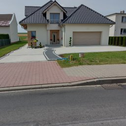 adamex-bruk - Nowoczesny dom jednorodzinny z podjazdem wyłożonym jasną kostką brukową z ciemnymi akcentami i fragmentem czerwonej kostki przy ulicy. Uwagę zwraca starannie zaprojektowany ogród.