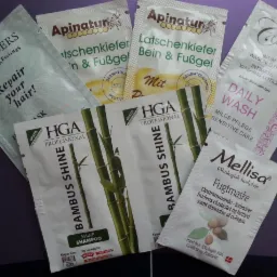 Zestaw próbek kosmetyków różnych marek, w tym maseczki do włosów, balsamy do ciała i szampony, ułożonych na fioletowym tle. Widoczne opakowania z nazwami firm: Stenders, Apinatur, HGA Professional,...