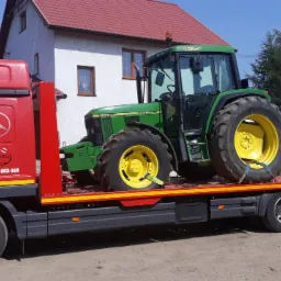 Czerwona laweta Mercedes Atego z załadowanym zielonym traktorem John Deere 6400, zabezpieczonym pasami transportowymi, na tle wiejskiego krajobrazu z domem i budynkiem gospodarczym.