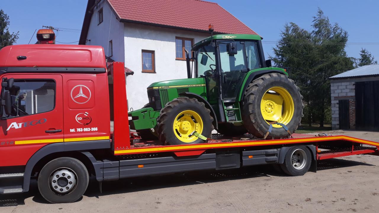 Czerwona laweta Mercedes Atego z załadowanym zielonym traktorem John Deere 6400, zabezpieczonym pasami transportowymi, na tle wiejskiego krajobrazu z domem i budynkiem gospodarczym.