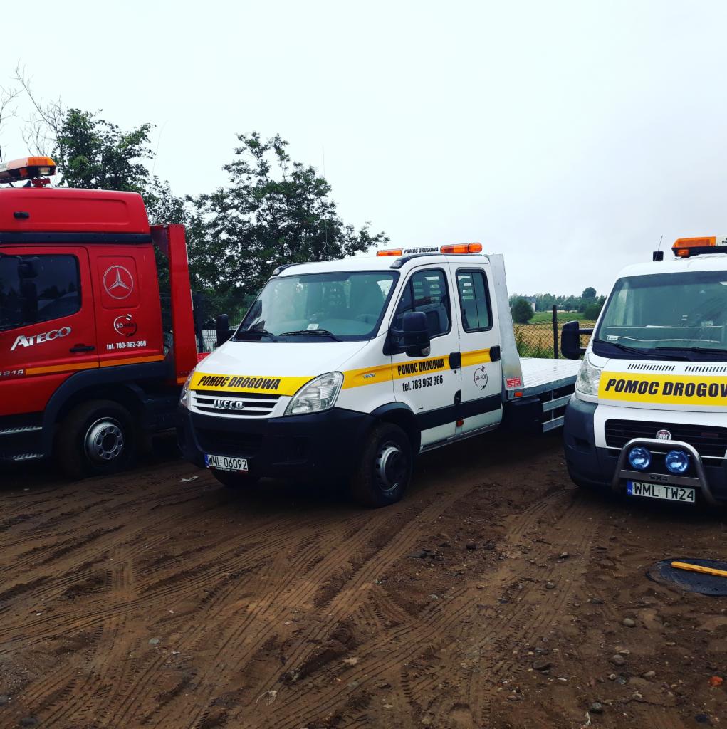Trzy pojazdy pomocy drogowej w błotnistym terenie, z widocznymi napisami 'POMOC DROGOWA' i numerami telefonów, różne modele Iveco i Atego z pomarańczowymi światłami ostrzegawczymi.