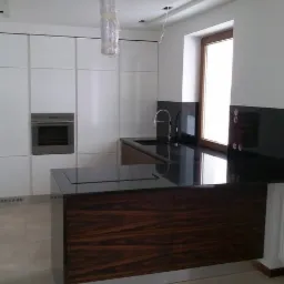 Nowoczesna kuchnia z białymi, gładkimi szafkami, wbudowanym piekarnikiem, wyspą z ciemnego drewna egzotycznego i czarnym blatem oraz czarnym panelem nad blatem przy oknie.