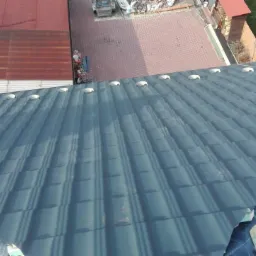 Montaż ciemnoszarej blachodachówki z widocznymi otworami wentylacyjnymi na membranie dachowej, w tle sąsiedni budynek z czerwoną blachą trapezową i kostka brukowa.