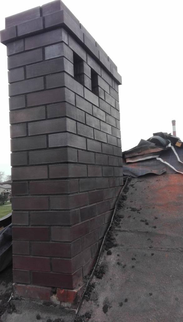 Murowany komin z cegły klinkierowej w kolorze ciemnego brązu, wystający ponad zniszczoną, bitumiczną powierzchnię dachu, z widocznymi śladami prac remontowych i fragmentami papy.