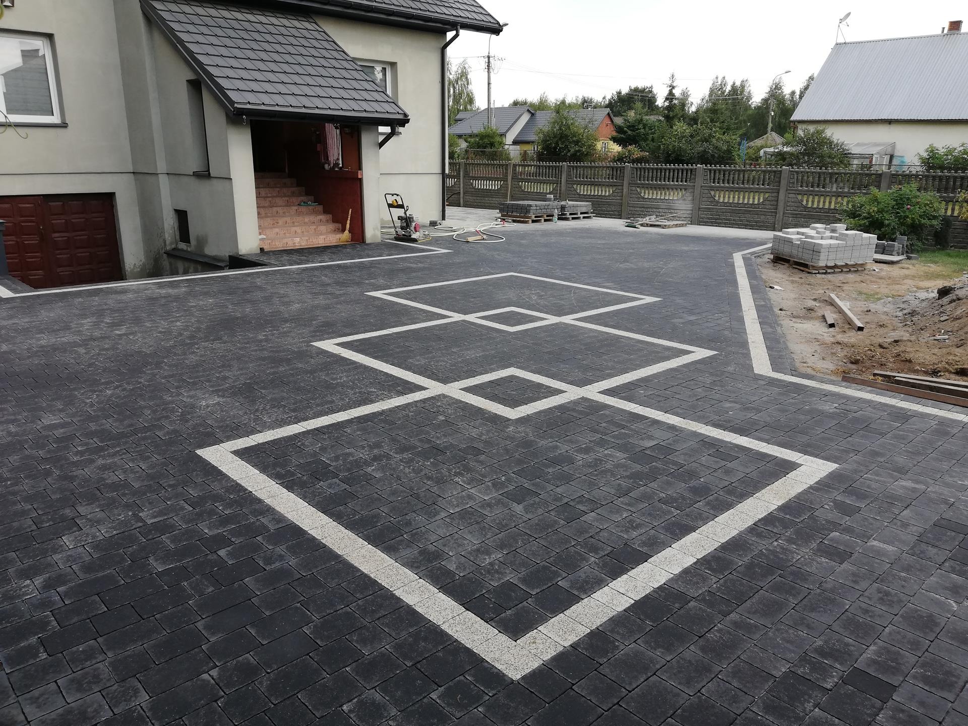 Ułożona kostka brukowa w Zalesiu z wzorem trzech połączonych kwadratów obramowanych jaśniejszą kostką, widoczny dom z garażem i palety z kostką. Przy domu stoi zagęszczarka.