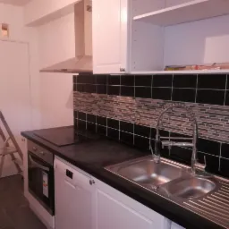 Wykończona kuchnia z czarnymi płytkami i mozaiką nad blatem, widoczny zlew dwukomorowy z nowoczesną baterią, białe szafki oraz drabina malarska oparta o ścianę.