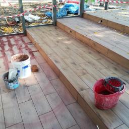 Darioremonty - Układanie drewnopodobnych płytek na tarasie zewnętrznym, widoczne wiadra z zaprawą i narzędzia na częściowo wykończonej powierzchni.