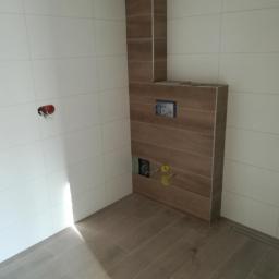 Darioremonty - Wnętrze łazienki w trakcie prac wykończeniowych, widoczne białe płytki na ścianach, zabudowa stelaża WC wykończona płytkami imitującymi drewno ułożonymi poziomo, oraz ułożone podłogowe panele...