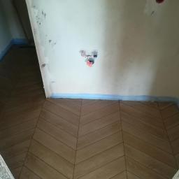 Darioremonty - Ułożona podłoga z płytek w jodełkę w trakcie remontu łazienki, ściany przygotowane do malowania, zabezpieczone niebieską taśmą malarską, widoczne wystające rury instalacji hydraulicznej...