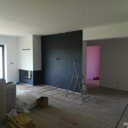 Darioremonty - Pokój w trakcie malowania z ciemną ścianą, zabezpieczoną podłogą, drabiną i kominkiem w tle, widoczne gniazdka elektryczne.