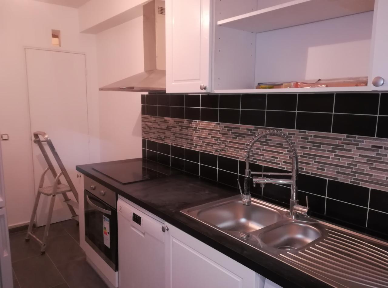 Wykończona kuchnia z czarnymi płytkami i mozaiką nad blatem, widoczny zlew dwukomorowy z nowoczesną baterią, białe szafki oraz drabina malarska oparta o ścianę.