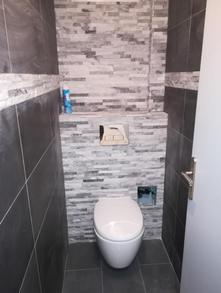 Wąska toaleta z szarymi płytkami imitującymi kamień na ścianach, podwieszana miska WC i ukryta spłuczka z chromowanym przyciskiem, widoczny niebieski odświeżacz powietrza na półce.