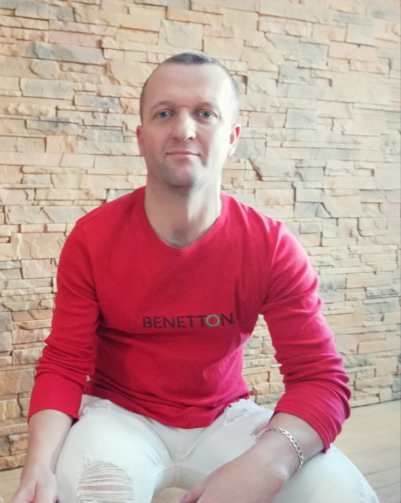 Mężczyzna w czerwonej koszulce Benetton siedzi na tle ściany z kamienia dekoracyjnego. Białe spodnie z dziurami, srebrna bransoletka na nadgarstku.