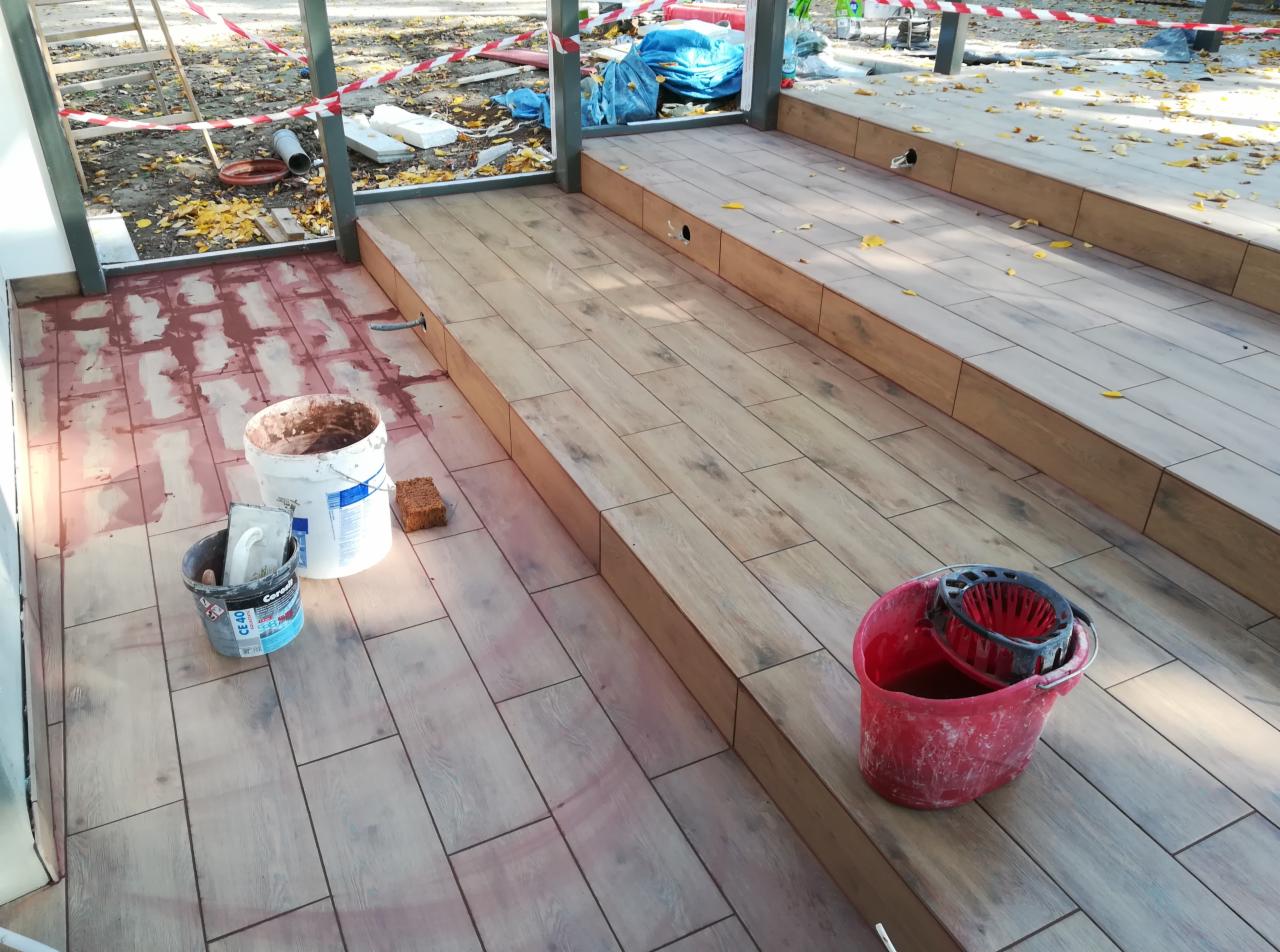 Układanie drewnopodobnych płytek na tarasie zewnętrznym, widoczne wiadra z zaprawą i narzędzia na częściowo wykończonej powierzchni.