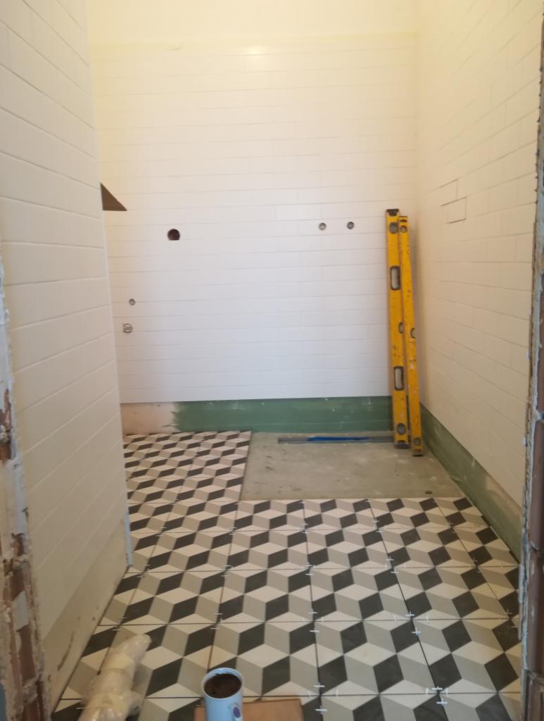 Łazienka w trakcie układania podłogi z płytek w geometryczny wzór 3D, poziomica oparta o ścianę z białej cegły, widoczne otwory pod instalacje.